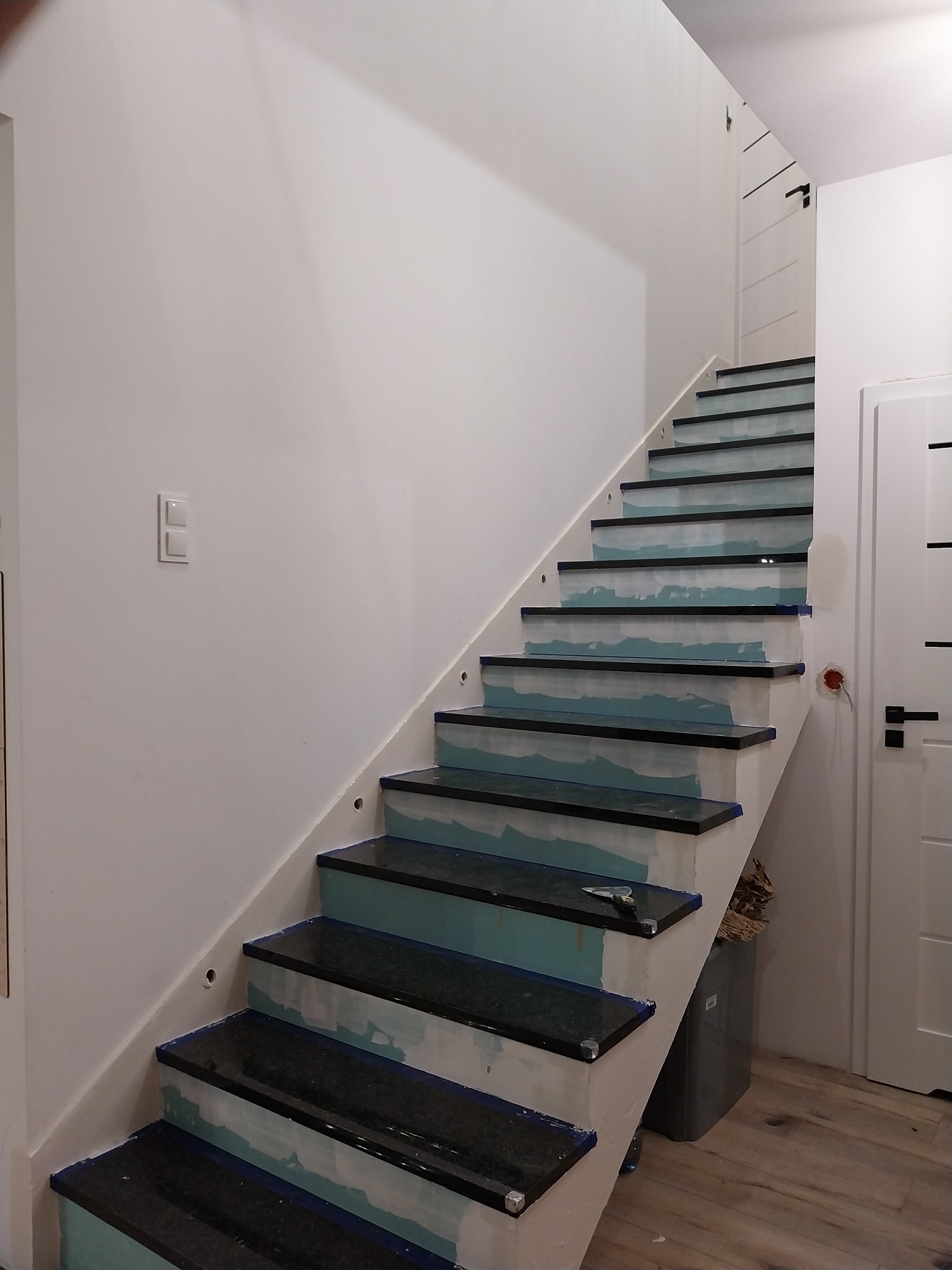 Montaż czarnych, granitowych stopni schodowych z niebieską taśmą zabezpieczającą, widoczne przygotowania do fugowania i szpachelka na jednym ze stopni.