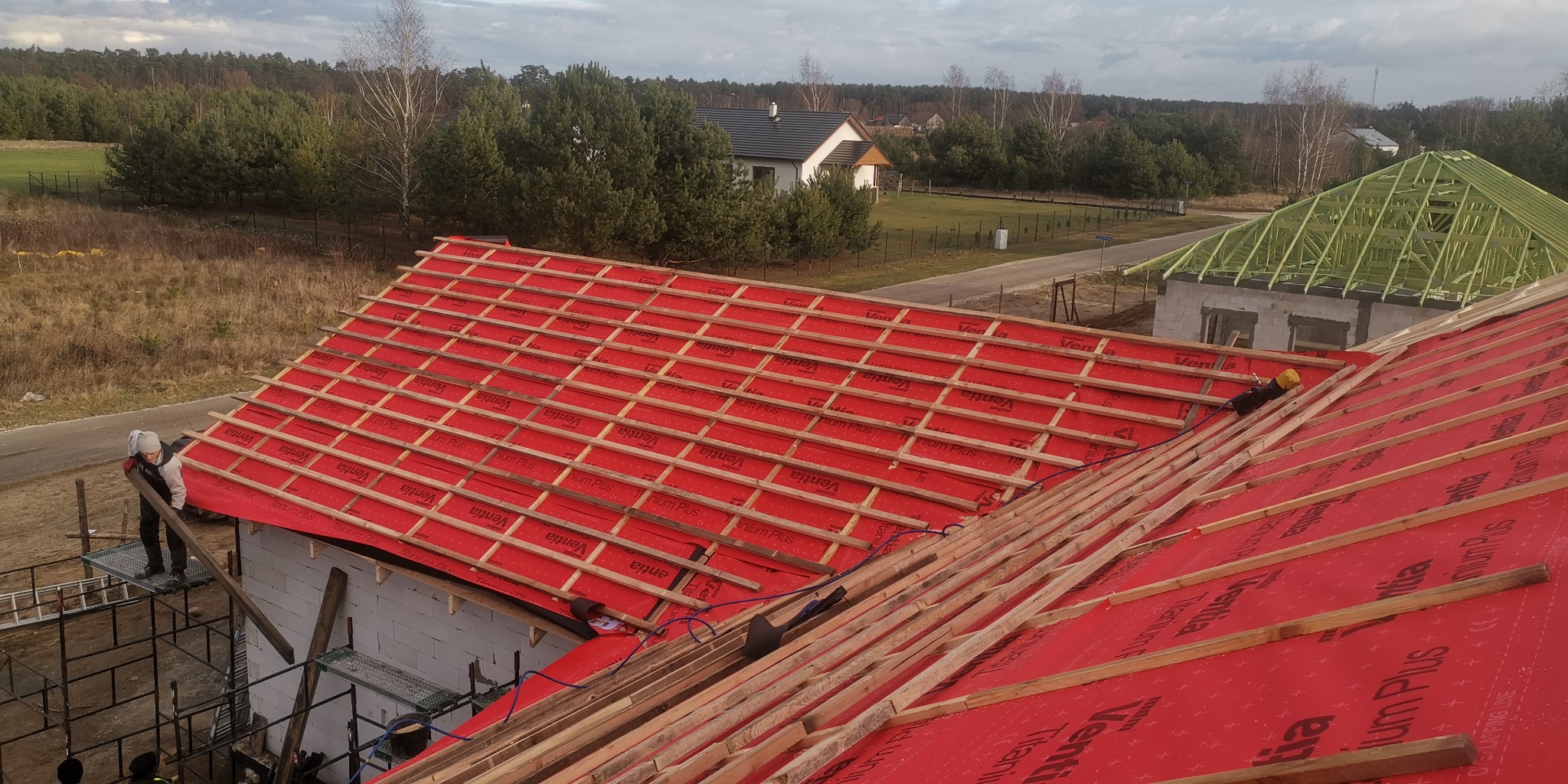 Montaż łat na czerwonej membranie dachowej, widoczni pracownicy na dachu i rusztowaniu, w tle domy jednorodzinne i drzewa.