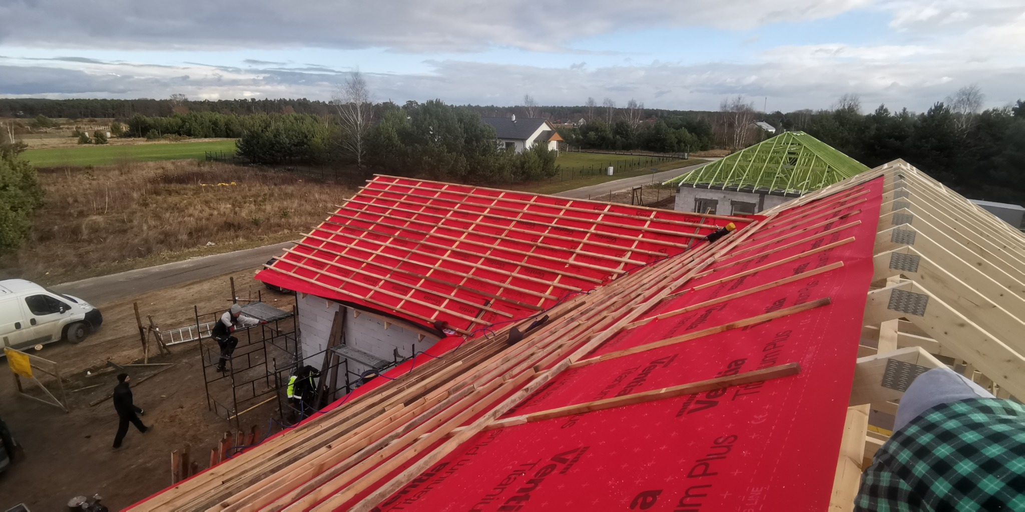 Montaż łat na czerwonej membranie dachowej na budowie domu jednorodzinnego, widok z góry na konstrukcję dachu i okoliczne domy.