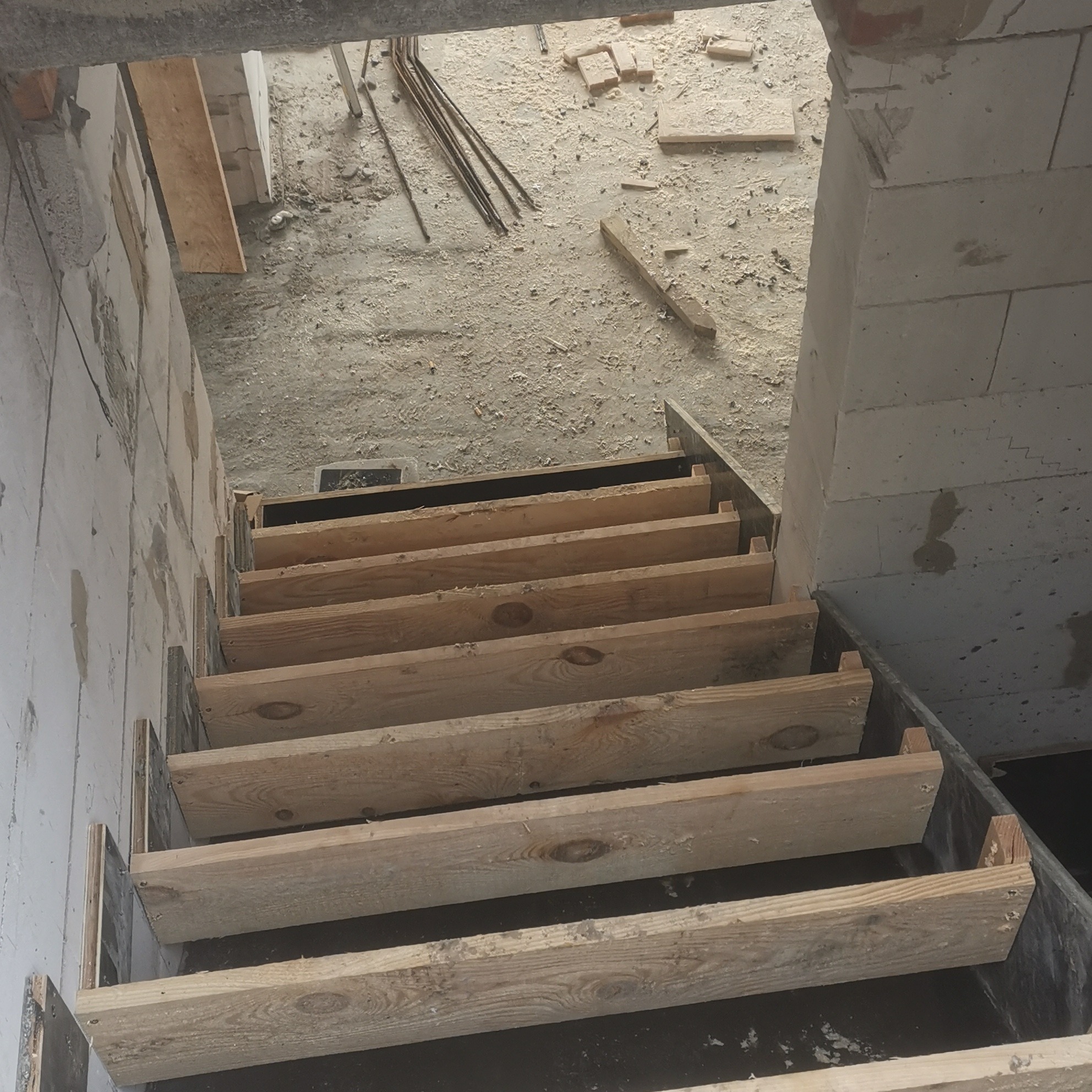 Surowe drewniane schody tymczasowe w surowym stanie, z widocznymi bloczkami betonowymi i gruzem budowlanym.