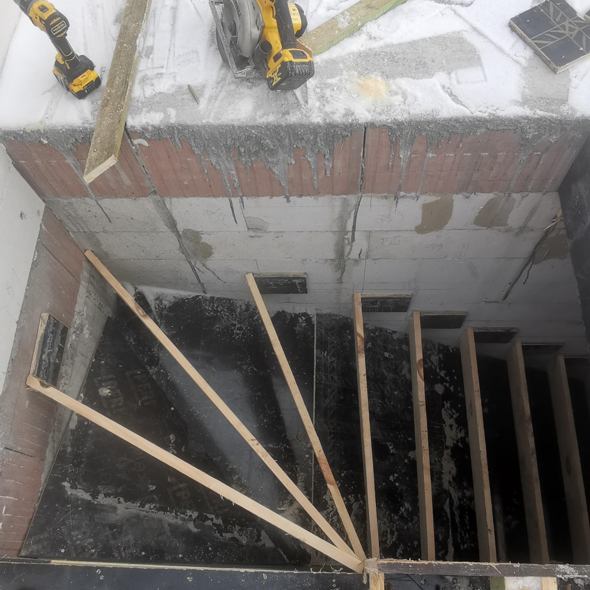 Wnętrze w trakcie budowy schodów z czarnymi płytami, widoczne drewniane elementy konstrukcyjne, wiertarka i piła tarczowa na górze.