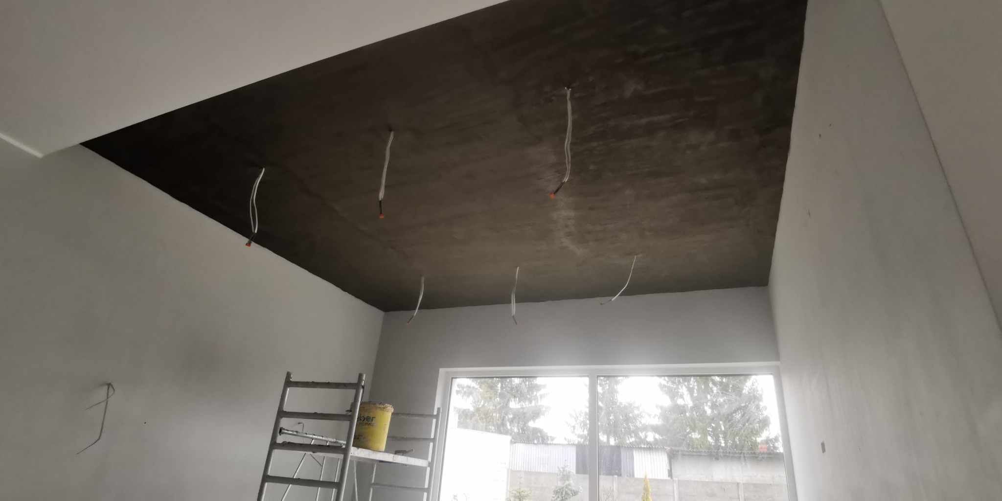 Szara surowa powierzchnia sufitu z wystającymi przewodami elektrycznymi w nowo wykończonym pomieszczeniu, drabina malarska i widok na zewnątrz przez okno.