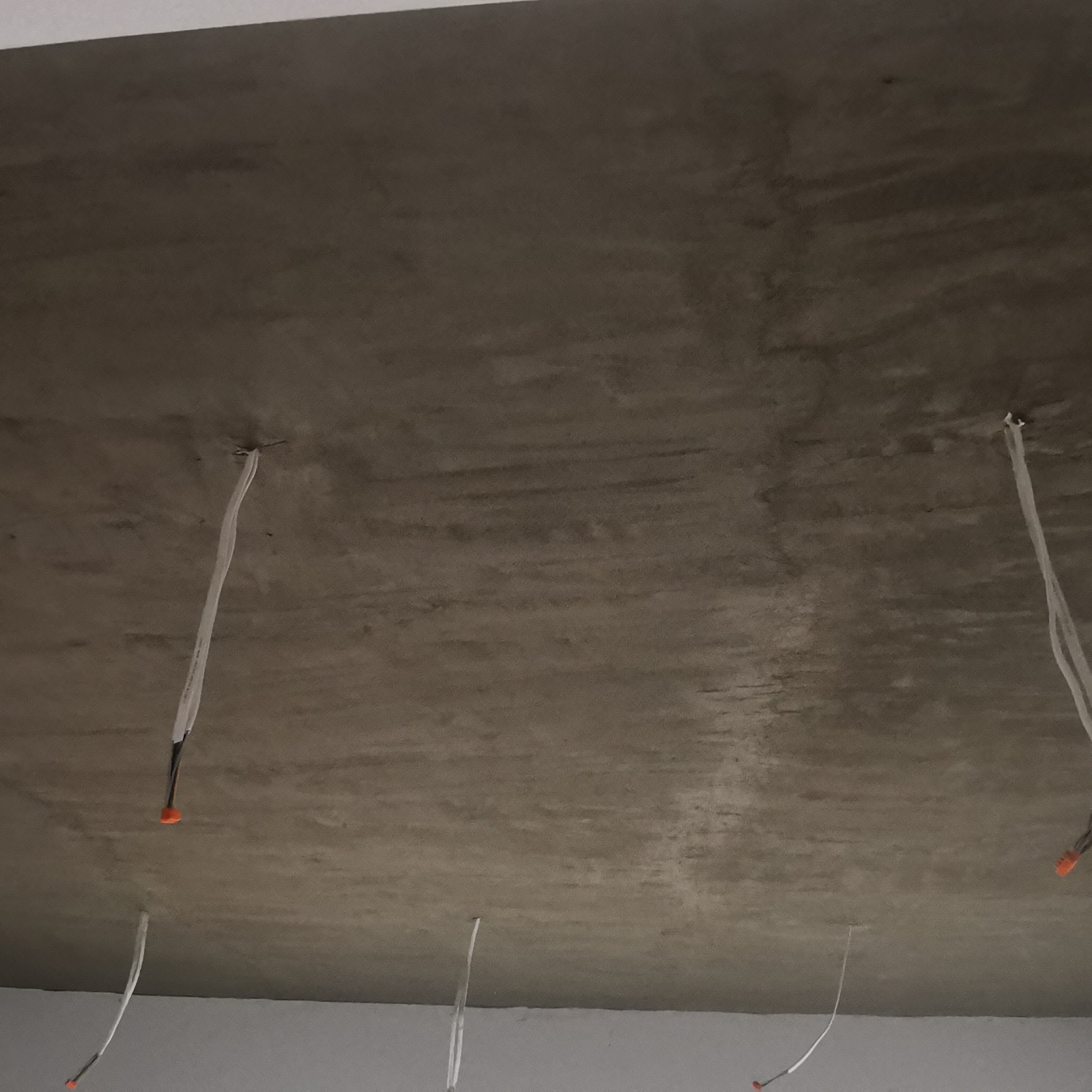 Surowy betonowy sufit z wystającymi przewodami elektrycznymi zabezpieczonymi pomarańczowymi złączkami, widoczne ślady po szalunku.