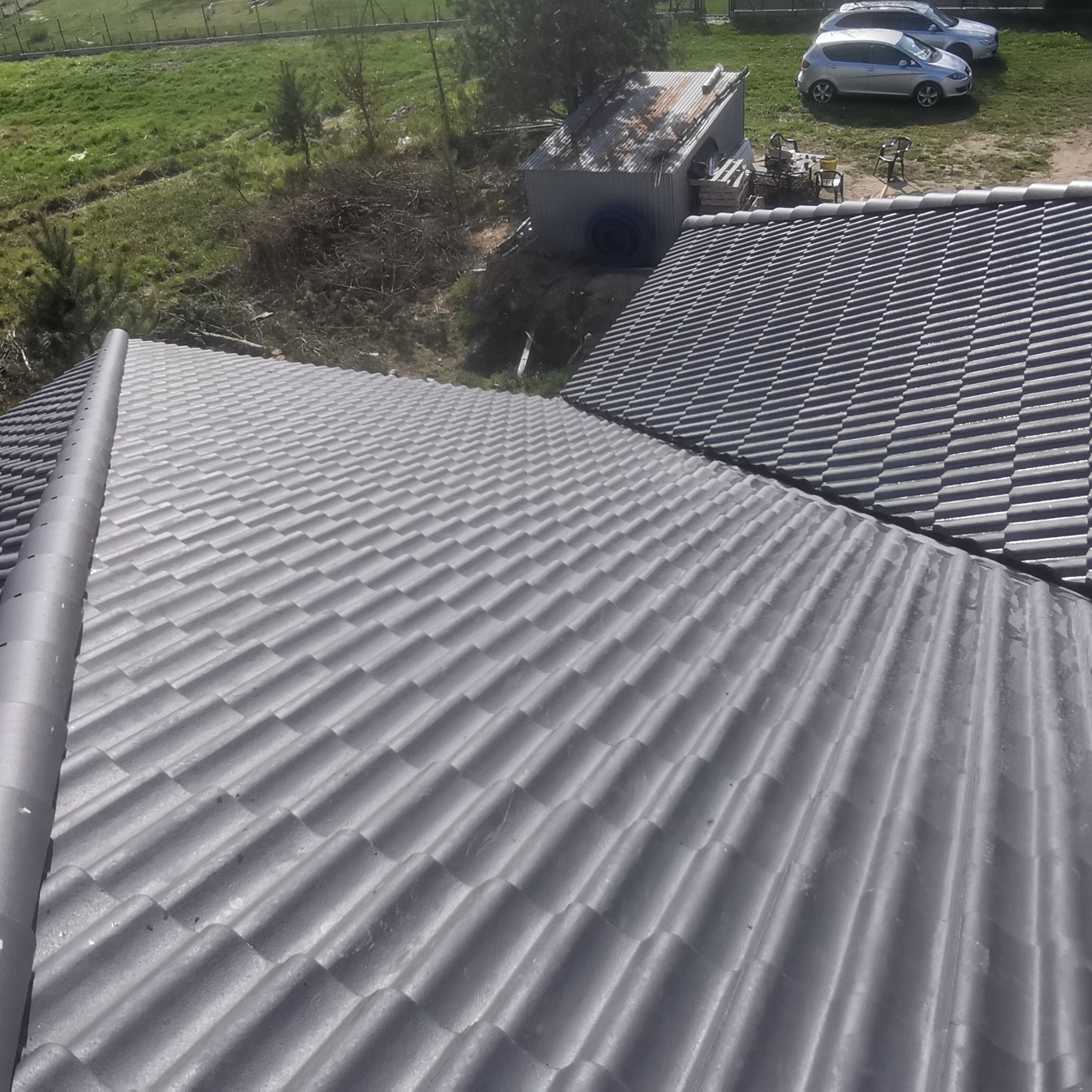 Widok z góry na fragment szarego dachu z dachówki ceramicznej, z rynną na krawędzi, na tle zabudowań gospodarczych, trawnika i zaparkowanych samochodów.