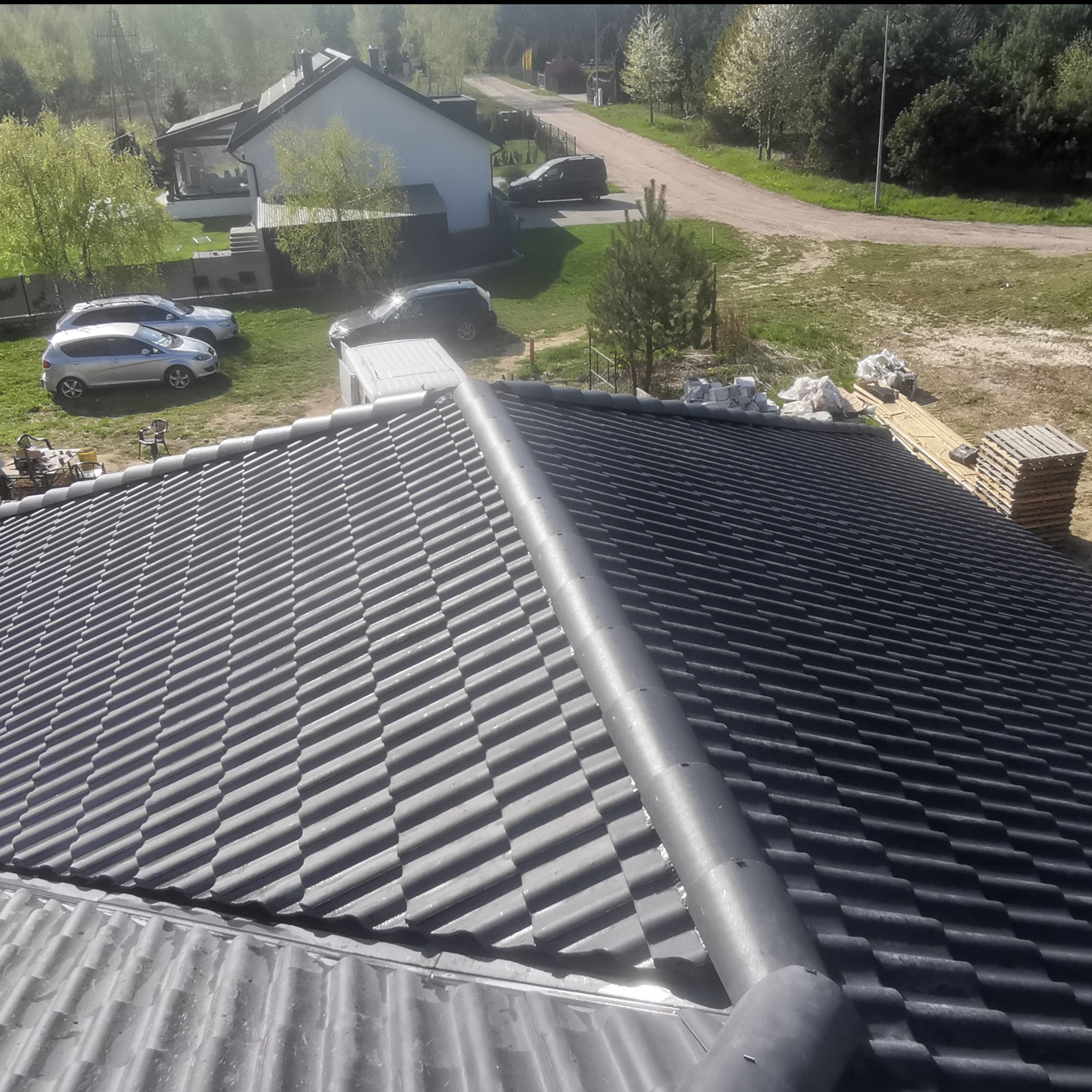 Nowy dach z ciemnoszarej dachówki ceramicznej, widok z góry na ukończoną połać, z widocznymi elementami wykończeniowymi i kominami wentylacyjnymi, w tle zabudowa jednorodzinna i zieleń.
