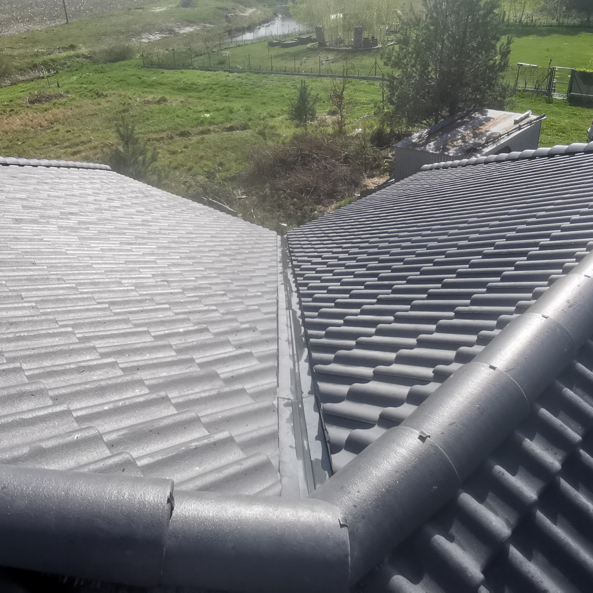 Szare dachówki ceramiczne na dachu domu w Bydgoszczy, widok z góry na kalenicę i system rynnowy, w tle zielony krajobraz.