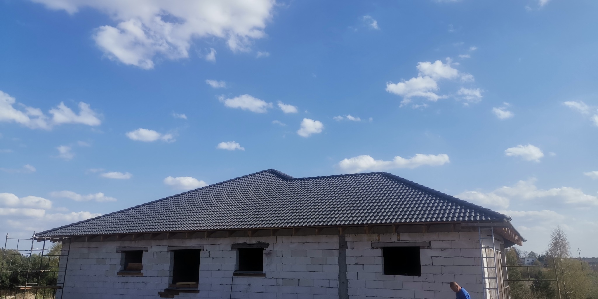 Dom murowany w trakcie budowy z widocznym dachem z ciemnej dachówki i ścianami z białego bloczka, na tle błękitnego nieba z chmurami.