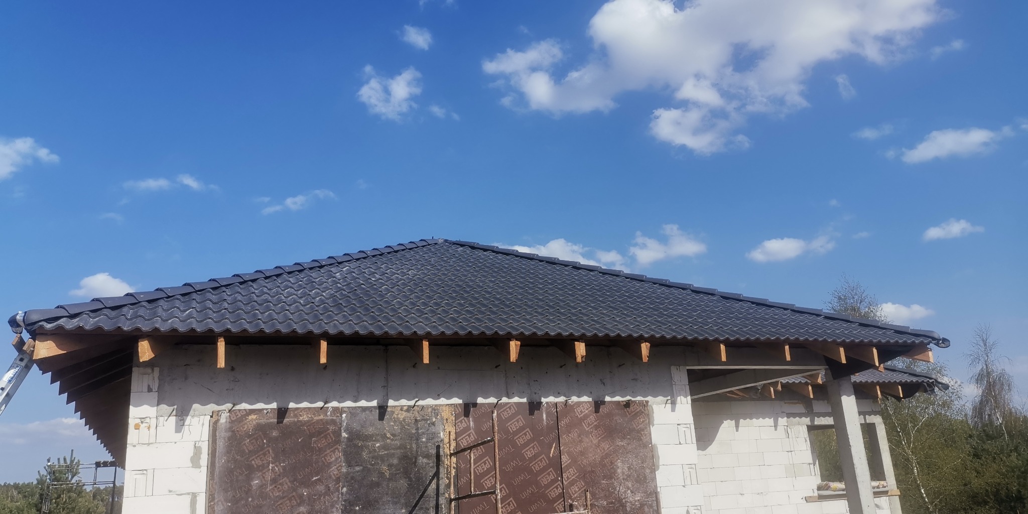 Budowa domu w trakcie realizacji: widok na fragment dachu z czarnej dachówki ceramicznej, ściany z białego betonu komórkowego częściowo zakryte płytami OSB, błękitne niebo z chmurami w tle.