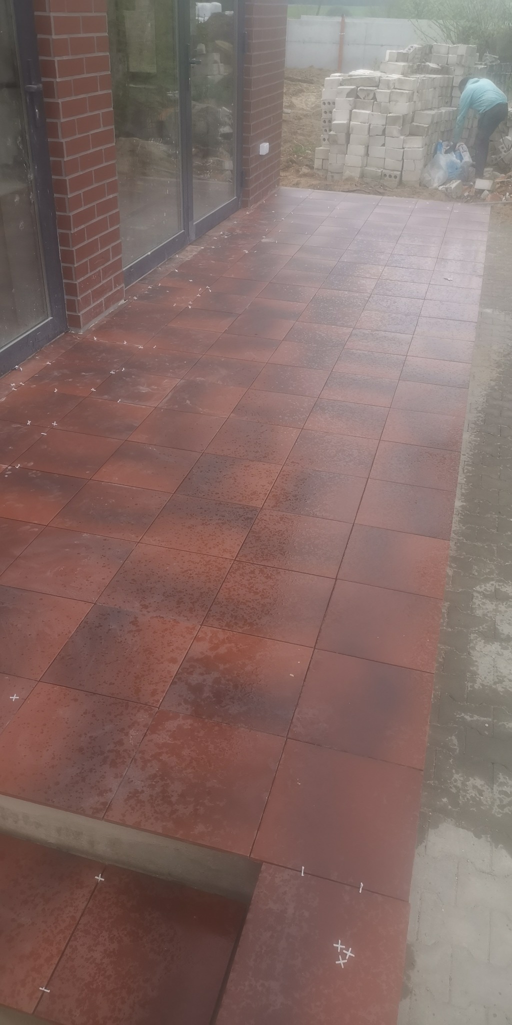 Ułożona, brązowa, kwadratowa terakota na tarasie przed budynkiem z ceglaną elewacją i dużymi oknami; w tle sterta bloczków budowlanych i pracownik w niebieskiej kurtce.