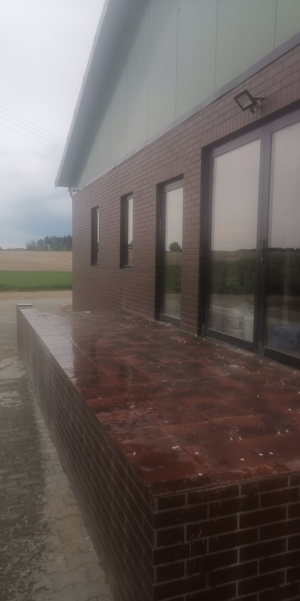 Nowoczesny budynek z cegły klinkierowej w kolorze brązowym, z dużymi oknami i wykończonym tarasem z płytek w podobnym odcieniu. Widoczny fragment elewacji z oświetleniem zewnętrznym oraz zielone...