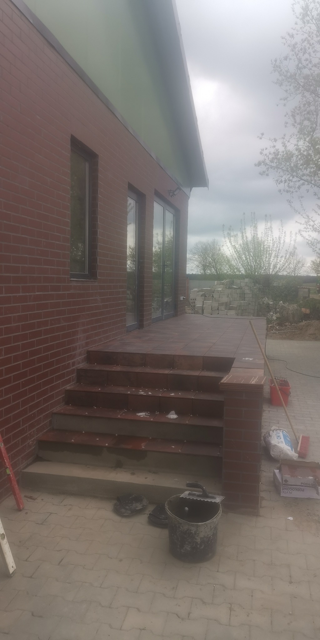 Wykończony taras z brązowych płytek przy ceglanym domu, widoczne schody wejściowe i narzędzia glazurnicze, pochmurne niebo w tle.