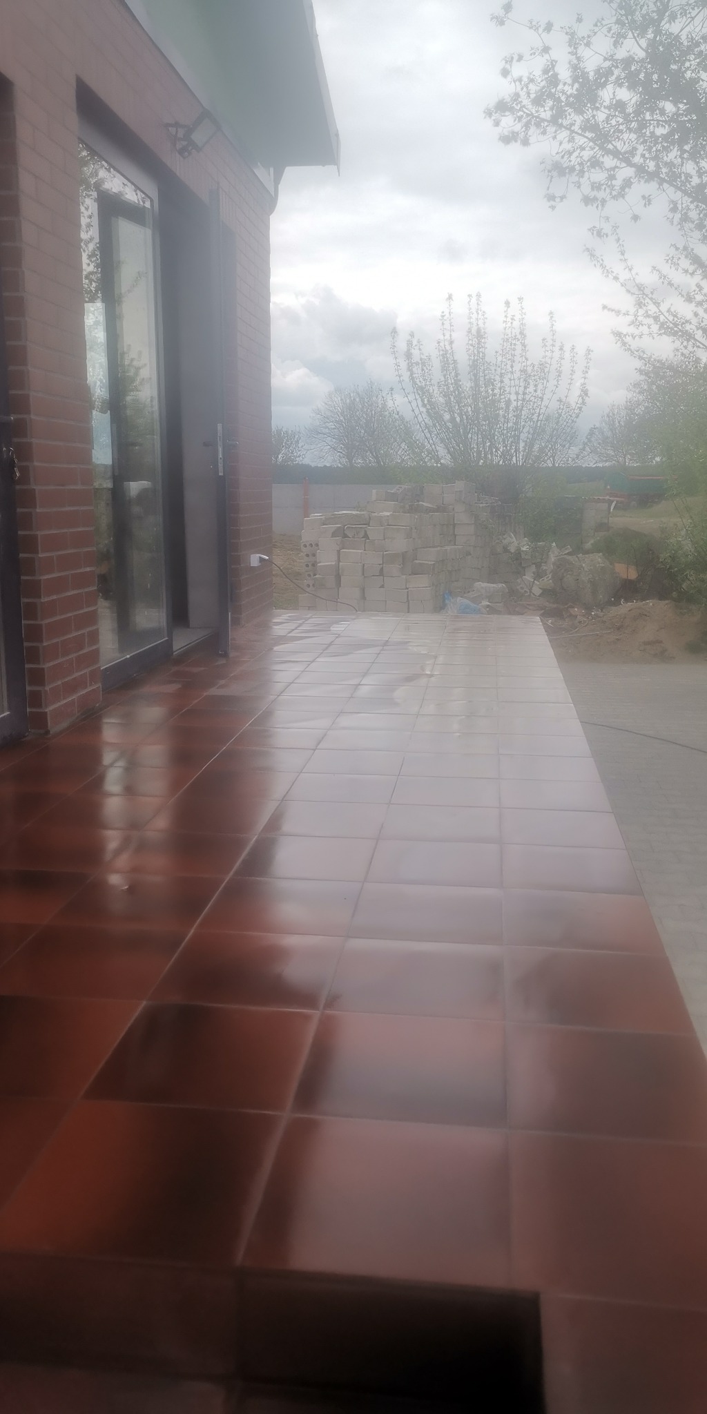 Błyszcząca, brązowa terakota ułożona na tarasie przy murowanym domu z widocznymi drzwiami balkonowymi i stertą bloczków betonowych w tle.