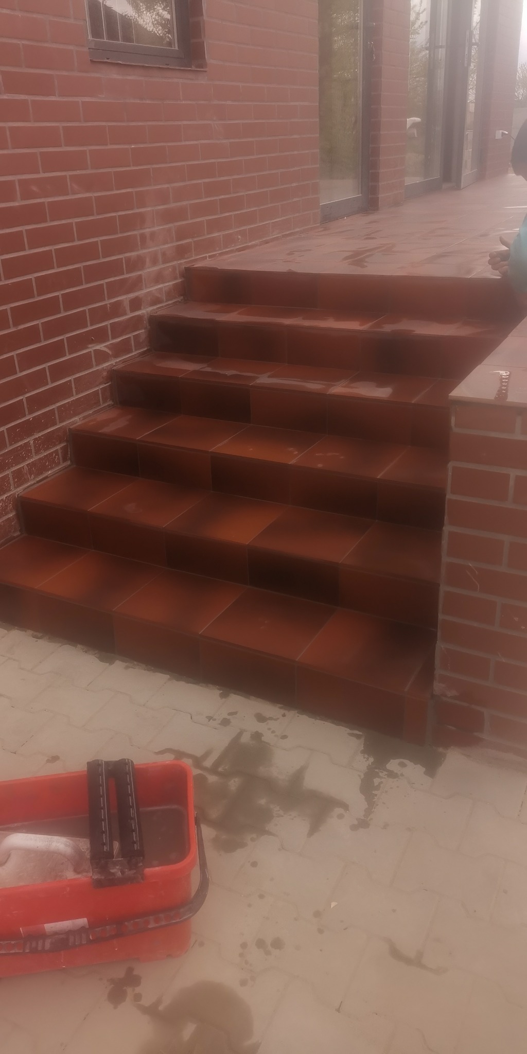 Wykończone płytkami schody wejściowe do domu z cegły klinkierowej, widoczny czerwony pojemnik z narzędziami glazurniczymi i mokra powierzchnia płytek.