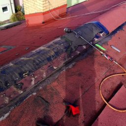 Mp-Flachdach Design - Czerwony dach pokryty papą z widocznymi śladami naprawy, narzędzia dekarskie i przewód gazowy leżące na powierzchni, w tle budynek mieszkalny.