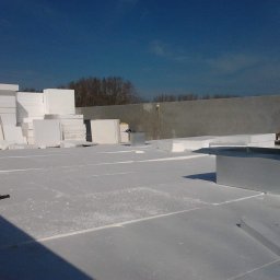 Mp-Flachdach Design - Białe płyty styropianowe ułożone na płaskim dachu, przygotowane do izolacji termicznej, z widocznym fragmentem ściany attyki w tle i błękitnym niebem.