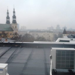 Mp-Flachdach Design - Widok z dachu w Poznaniu, na pierwszym planie płaska powierzchnia dachu pokryta papą, dwa klimatyzatory marki Samsung, w tle panorama miasta spowita mgłą, widoczne wieże kościoła.