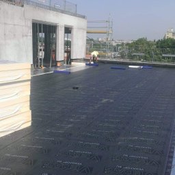 Mp-Flachdach Design - Prace na dachu budynku w trakcie budowy: dekarz w żółtej kamizelce układa czarną papę z napisem Vedag, obok stos izolacji termicznej w białej folii, w tle widok na miasto.