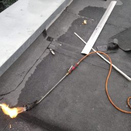 Mp-Flachdach Design - Prace dekarskie na dachu płaskim: palnik dekarski w użyciu, widoczna spoina, narzędzia dekarskie i fragment komina w tle.