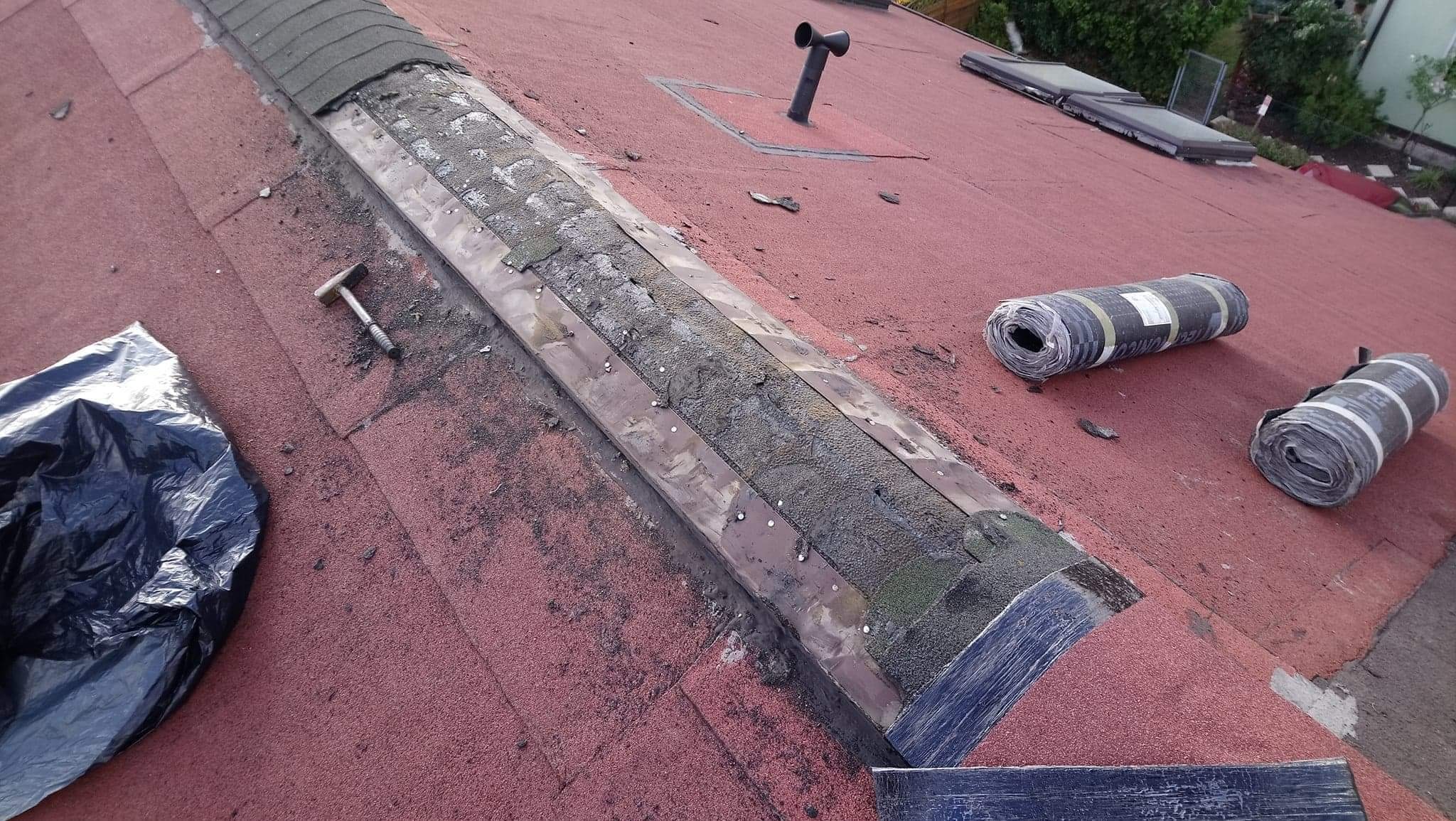 Czerwony dach pokryty papą z widocznym uszkodzonym fragmentem, narzędziem (młotek), workiem na odpady i rolkami papy bitumicznej w tle.