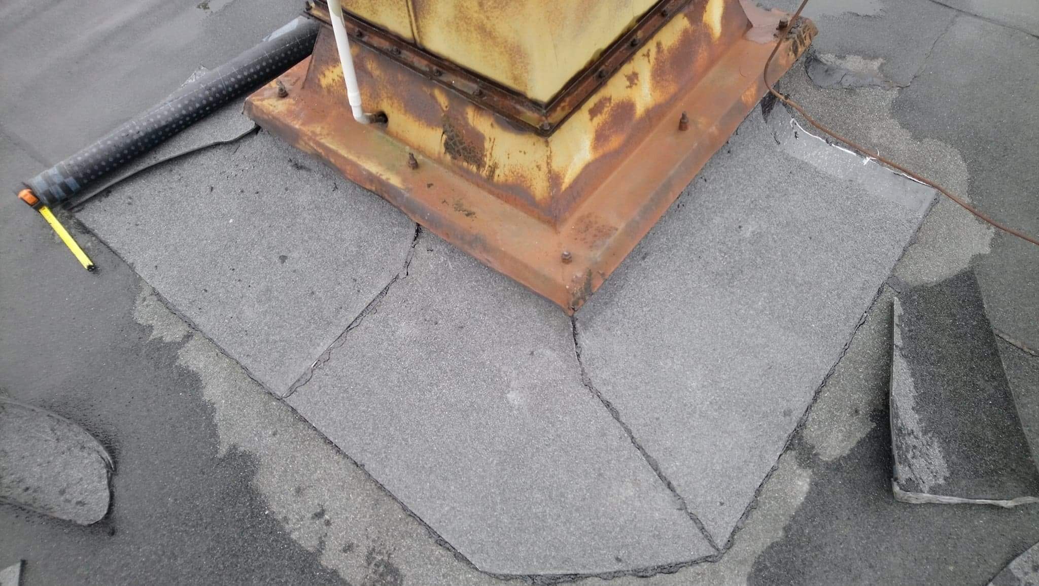 Płaski dach pokryty papą wokół zardzewiałej podstawy komina wentylacyjnego, widoczne ślady napraw i uszczelniania, obok leży zwinięta rolka papy i miarka.