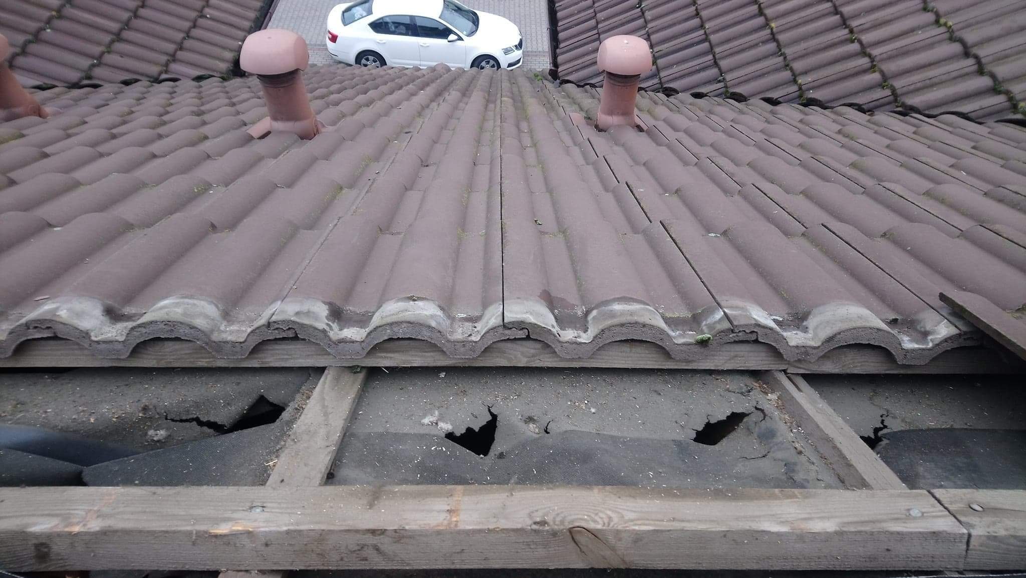 Uszkodzony dach z brązowej dachówki ceramicznej, widoczne dziury w membranie i elementy więźby dachowej, w tle biały samochód osobowy.