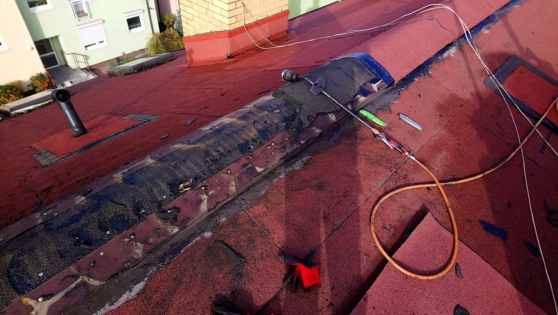 Czerwony dach pokryty papą z widocznymi śladami naprawy, narzędzia dekarskie i przewód gazowy leżące na powierzchni, w tle budynek mieszkalny.