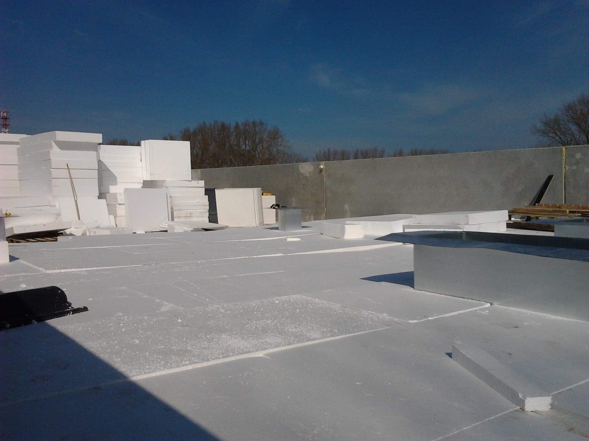 Białe płyty styropianowe ułożone na płaskim dachu, przygotowane do izolacji termicznej, z widocznym fragmentem ściany attyki w tle i błękitnym niebem.