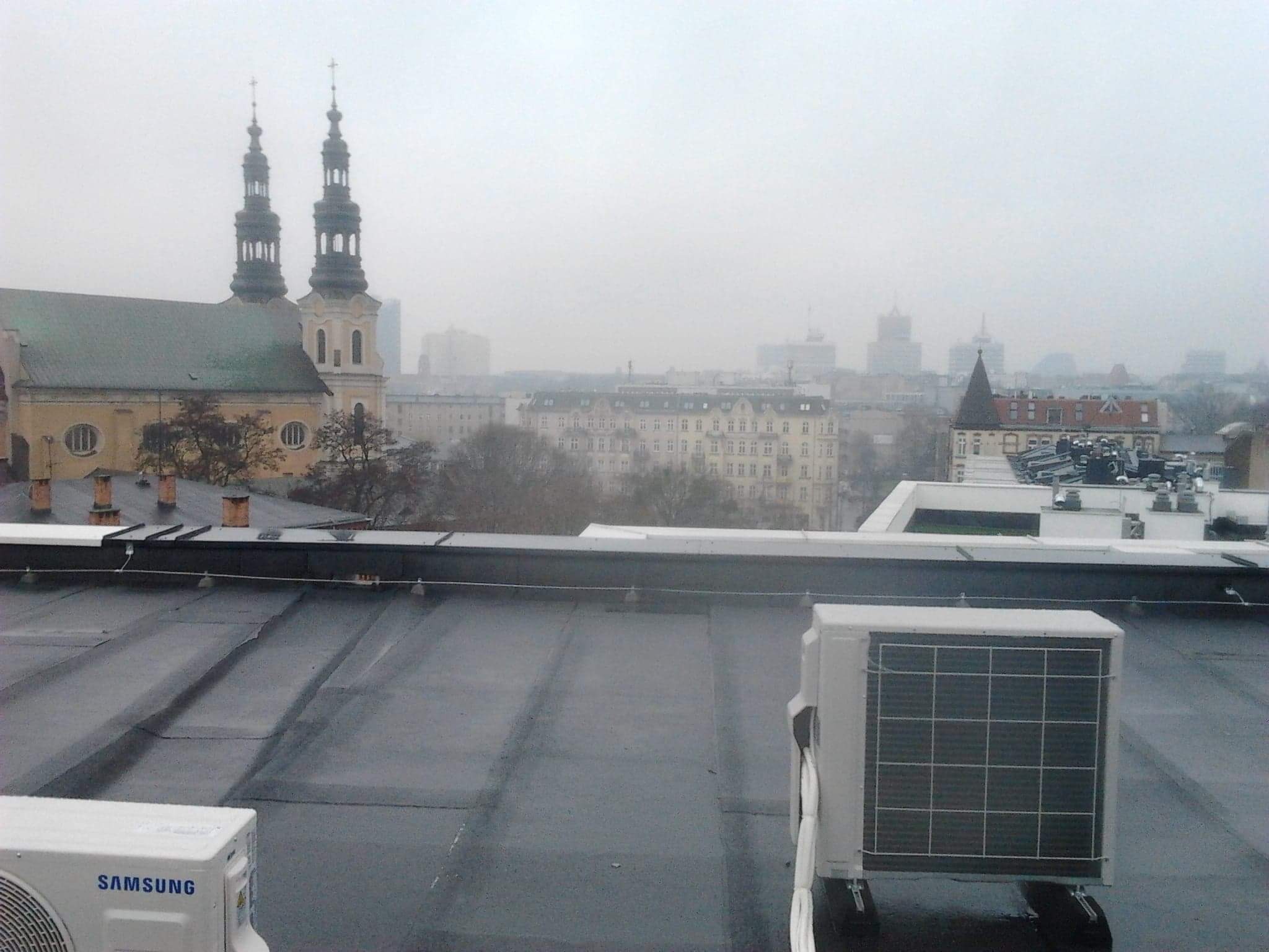 Widok z dachu w Poznaniu, na pierwszym planie płaska powierzchnia dachu pokryta papą, dwa klimatyzatory marki Samsung, w tle panorama miasta spowita mgłą, widoczne wieże kościoła.