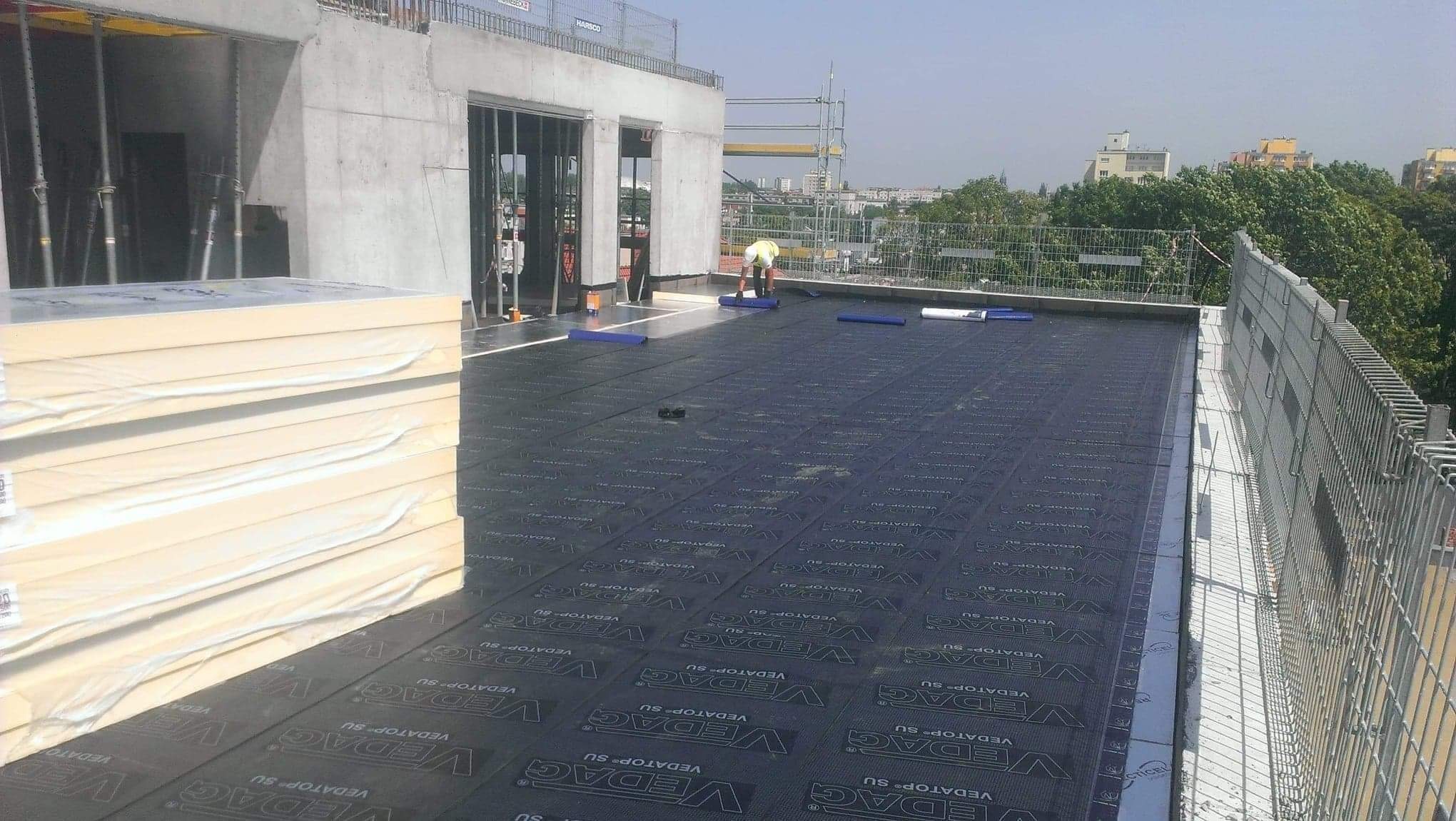 Prace na dachu budynku w trakcie budowy: dekarz w żółtej kamizelce układa czarną papę z napisem Vedag, obok stos izolacji termicznej w białej folii, w tle widok na miasto.