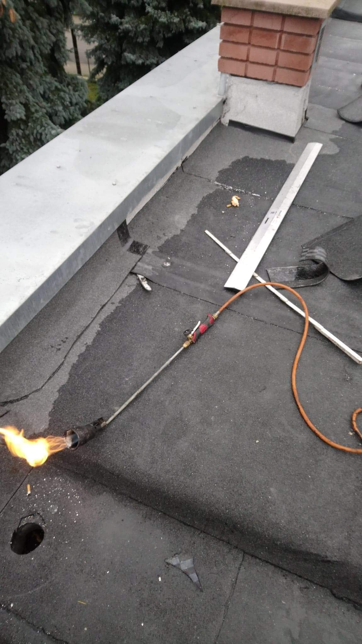 Prace dekarskie na dachu płaskim: palnik dekarski w użyciu, widoczna spoina, narzędzia dekarskie i fragment komina w tle.