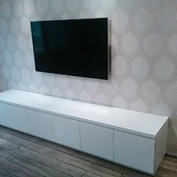 Długa, biała szafka RTV z połyskiem, zamontowana pod wiszącym telewizorem na ścianie z wzorzystą tapetą; widoczna drewniana podłoga.