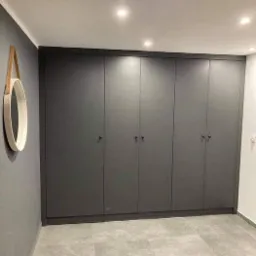 Szara zabudowa szafowa z sześcioma drzwiami, dopasowana do ściany, z okrągłym lustrem z drewnianym paskiem wiszącym obok, na tle szarej podłogi z płytek.