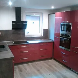 Nowoczesna kuchnia z bordowymi, lakierowanymi frontami szafek, szarym blatem i czarnym okapem nad płytą indukcyjną, z widoczną zabudową piekarnika i mikrofalówki.