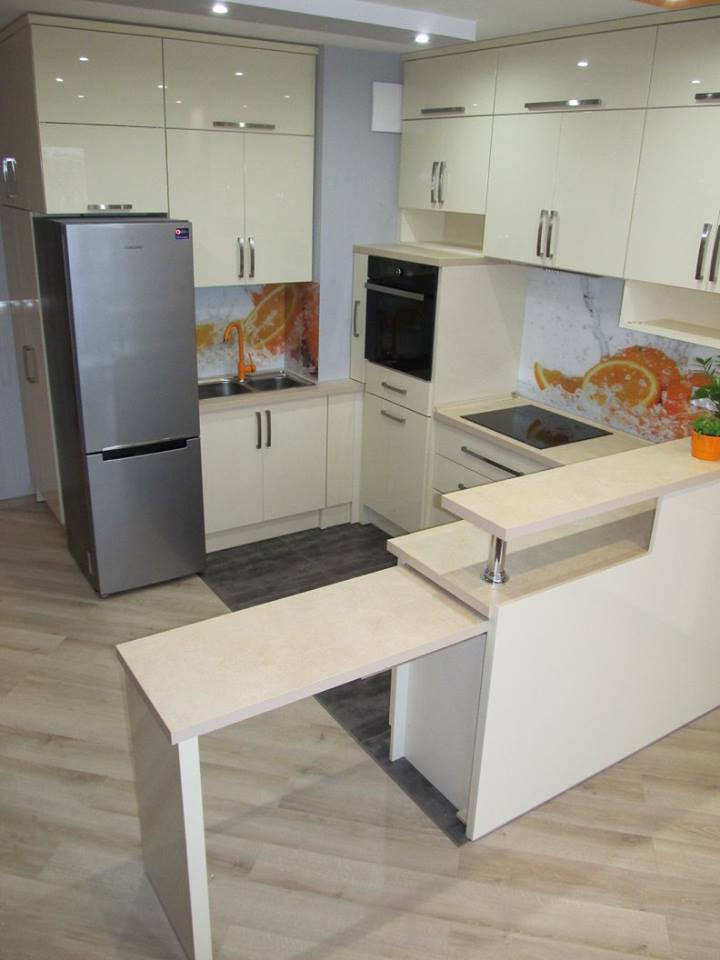 Jasna kuchnia z połyskiem, zabudowa na wymiar z blatem roboczym i wysuwanym blatem, srebrna lodówka i pomarańczowy motyw na ścianie.