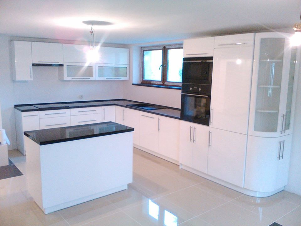 Nowoczesna, biała kuchnia z czarnymi blatami i wyspą, widoczna zabudowa piekarnika i przeszklona witryna narożna, jasna podłoga z płytek.