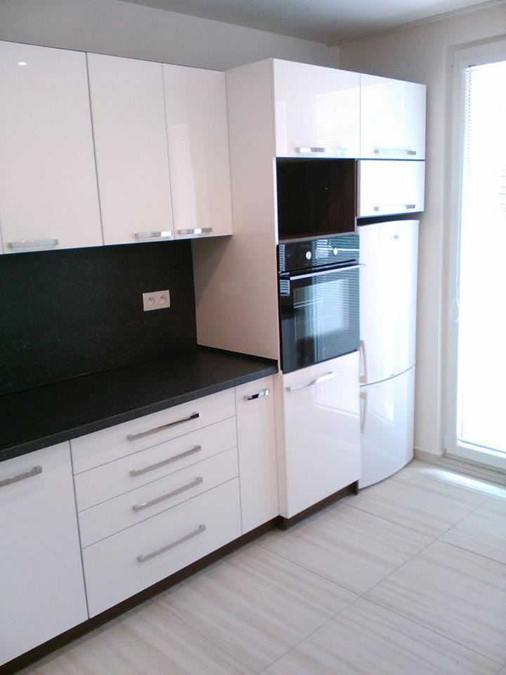 Biała, nowoczesna zabudowa kuchenna z czarnym blatem, srebrnymi uchwytami i wbudowanym piekarnikiem, obok lodówka i okno.