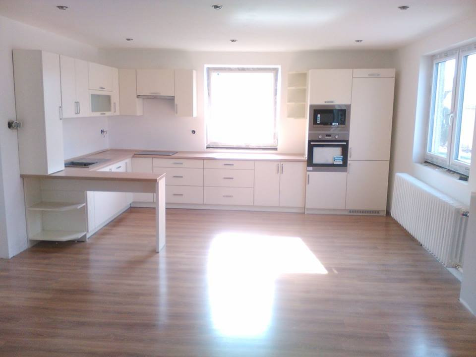 Jasna, nowoczesna kuchnia z białymi frontami, drewnianym blatem i półwyspem, oświetlona światłem wpadającym przez okno, z widocznym parkietem na podłodze.