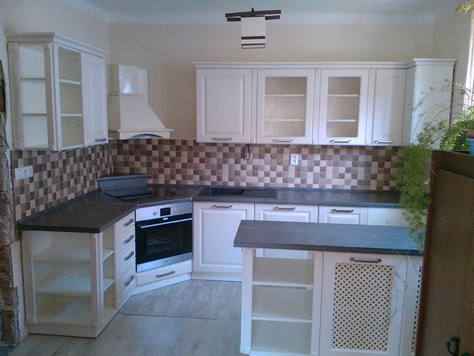 Jasna kuchnia z białymi szafkami i ciemnym blatem, mozaikowa ściana nad blatem, wyspa kuchenna z ażurowymi drzwiczkami.