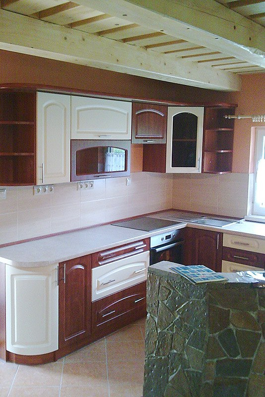 Kuchenna zabudowa w odcieniach brązu i beżu, z blatem o zaokrąglonym narożniku, widoczna wyspa wykończona kamieniem i surowy drewniany strop