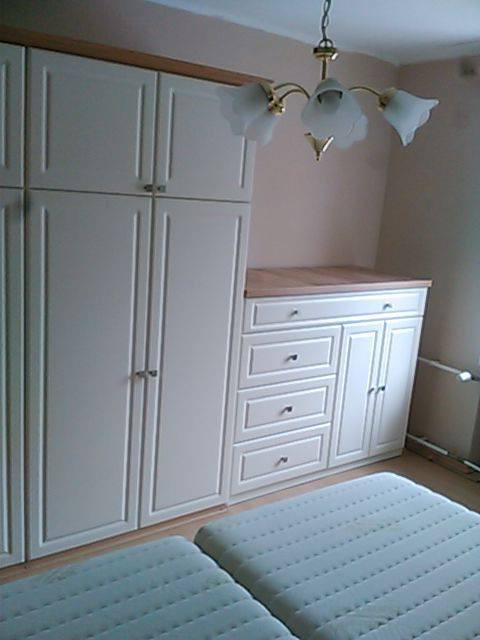 Zabudowa wnęki w sypialni z białymi frontami, obejmująca szafę i komodę z drewnianym blatem, oświetlona przez wiszącą lampę z białymi kloszami.