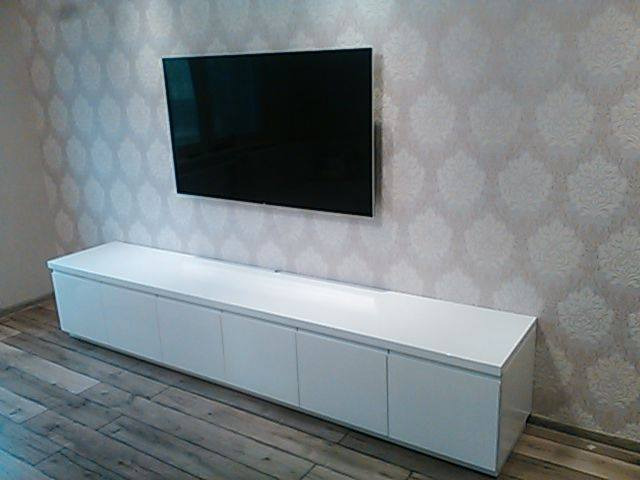 Długa, biała szafka RTV z połyskiem, zamontowana pod wiszącym telewizorem na ścianie z wzorzystą tapetą; widoczna drewniana podłoga.