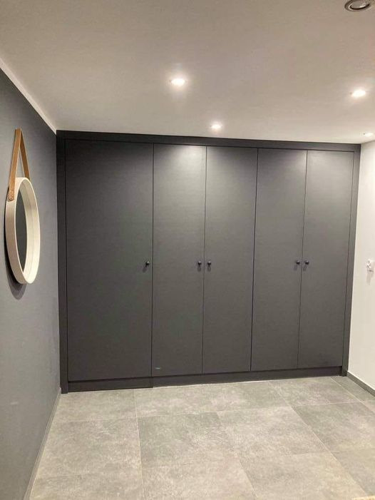 Szara zabudowa szafowa z sześcioma drzwiami, dopasowana do ściany, z okrągłym lustrem z drewnianym paskiem wiszącym obok, na tle szarej podłogi z płytek.