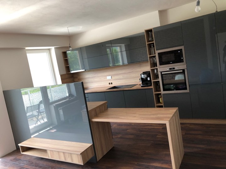 Nowoczesna kuchnia z szarymi, lakierowanymi frontami, drewnianym blatem i wyspą z przedłużonym blatem, widok na balkon przez odbicie w froncie wyspy.