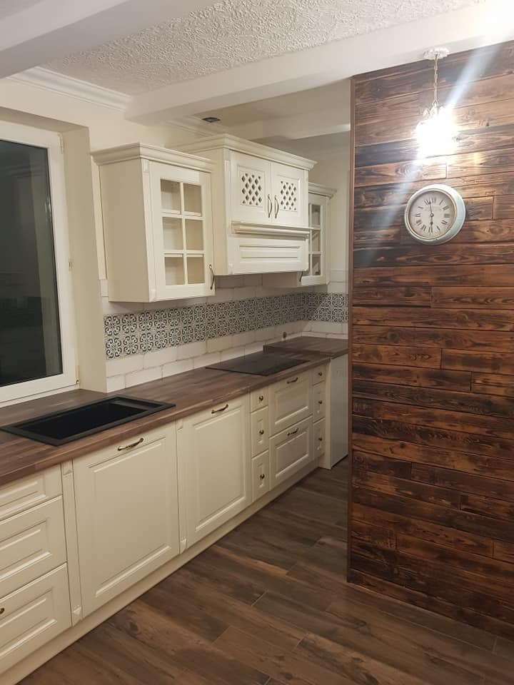 Jasna kuchnia z drewnianymi blatami, ozdobnymi kaflami nad blatem i ścianą wykończoną ciemnym drewnem z zegarem.