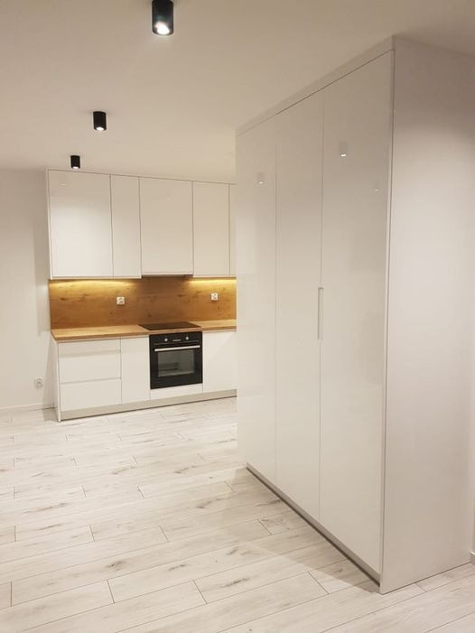 Nowoczesna kuchnia z białymi, lakierowanymi frontami szafek, drewnianym blatem i podświetleniem LED, uzupełniona wysoką szafą w zabudowie i jasną podłogą imitującą drewno.