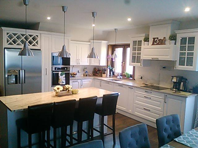 Jasna kuchnia w stylu klasycznym z wyspą i trzema lampami wiszącymi, zabudowa z szafkami z przeszklonymi frontami i wbudowanym regałem na wino nad lodówką.