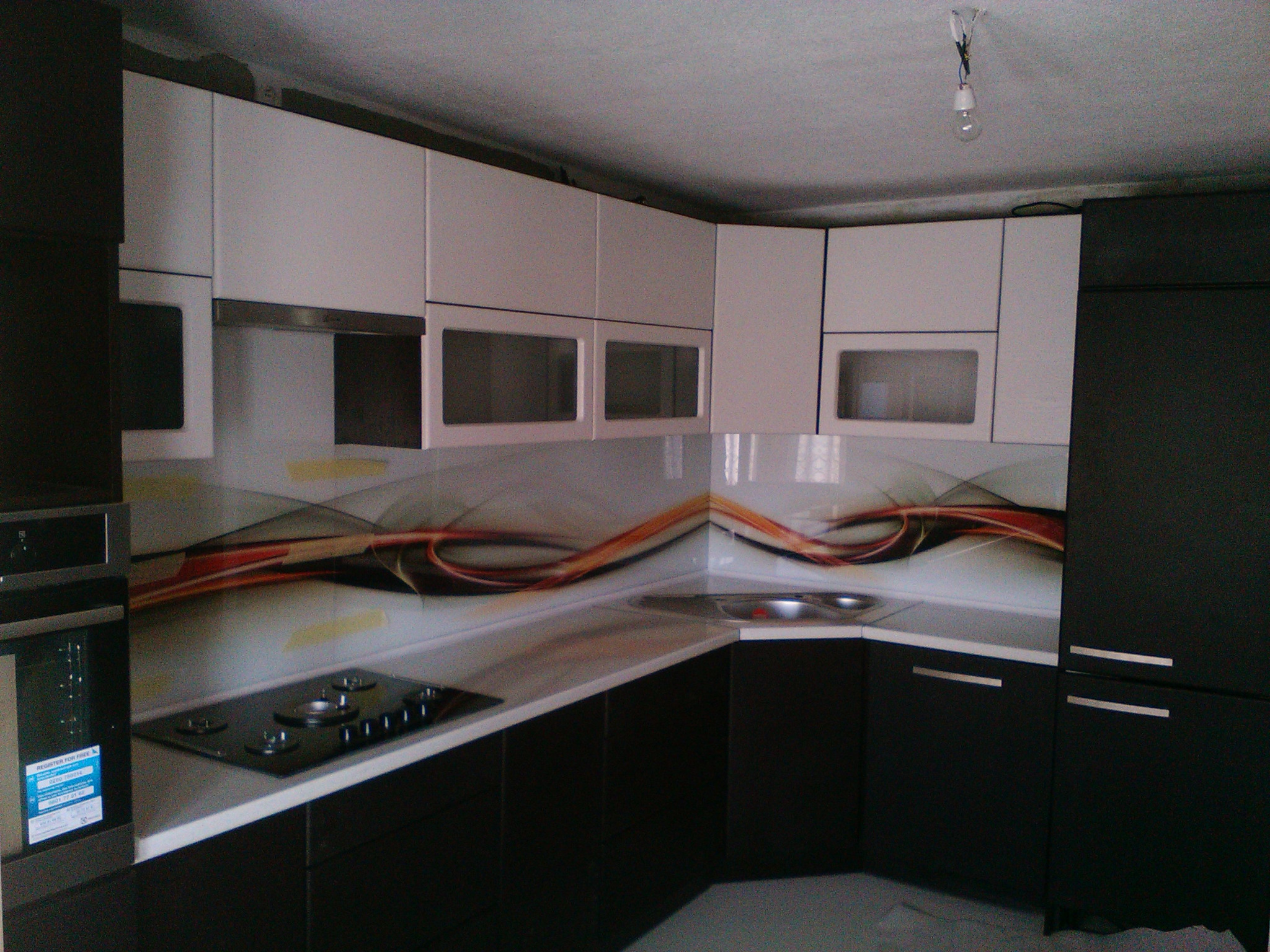 Nowoczesna kuchnia z czarnymi dolnymi szafkami i białymi górnymi, szklany panel nad blatem z abstrakcyjnym wzorem, płyta grzewcza i piekarnik w zabudowie, surowy stan wykończenia sufitu.