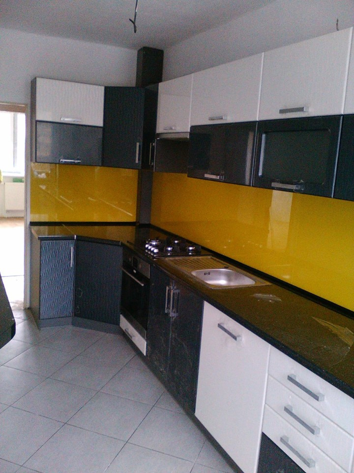 Nowoczesna kuchnia z żółtym panelem szklanym nad blatem, biało-czarne szafki z metalowymi uchwytami, widoczne ślady po remoncie na podłodze i szafkach.