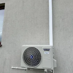 Zewnętrzna jednostka klimatyzatora Kaisai ICE/ICE BLACK zamontowana na szarej, teksturowanej ścianie budynku z widocznymi rurami odprowadzającymi skropliny i osłoną instalacji elektrycznej.