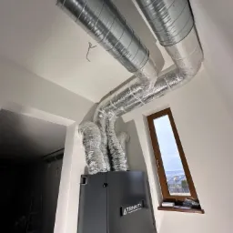 Czarna centrala wentylacyjna z rekuperacją marki Trinnity, z widocznymi przewodami wentylacyjnymi w folii aluminiowej, zamontowana w pomieszczeniu ze skośnym oknem i drabinką w tle.